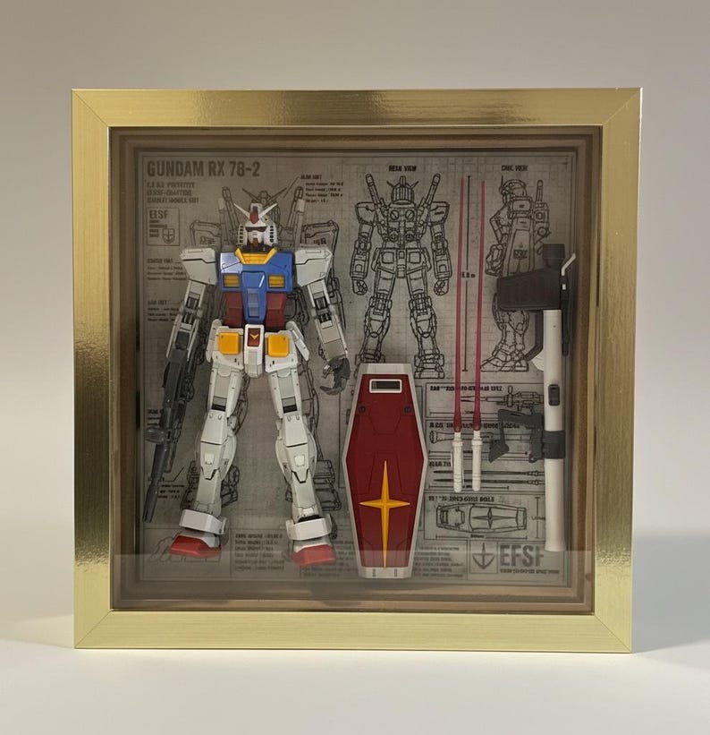 May include: A framed Gundam RX 78-2 model kit. The robot is white, blue, red, and yellow. Included are a red shield with a yellow cross, two red beam sabers, and a rifle. The background features blueprints.