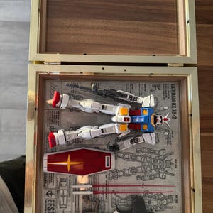 May include: A framed Gundam RX-78-2 model kit, featuring white, blue, red, and yellow components, along with a red shield with a yellow star. The model is displayed on a blueprint background within a gold-framed shadow box.