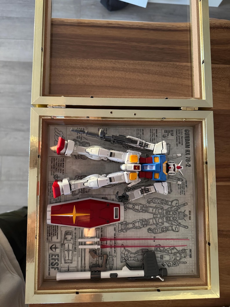 May include: A framed Gundam RX-78-2 model kit, featuring white, blue, red, and yellow components, along with a red shield with a yellow star. The model is displayed on a blueprint background within a gold-framed shadow box.