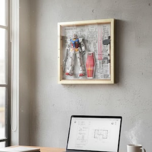 May include: A framed Gundam model kit displayed on a gray wall. The model is in a clear box with a wooden frame, showcasing the robot, shield, and blueprints. A laptop and a mug of coffee sit on a wooden desk in the foreground.