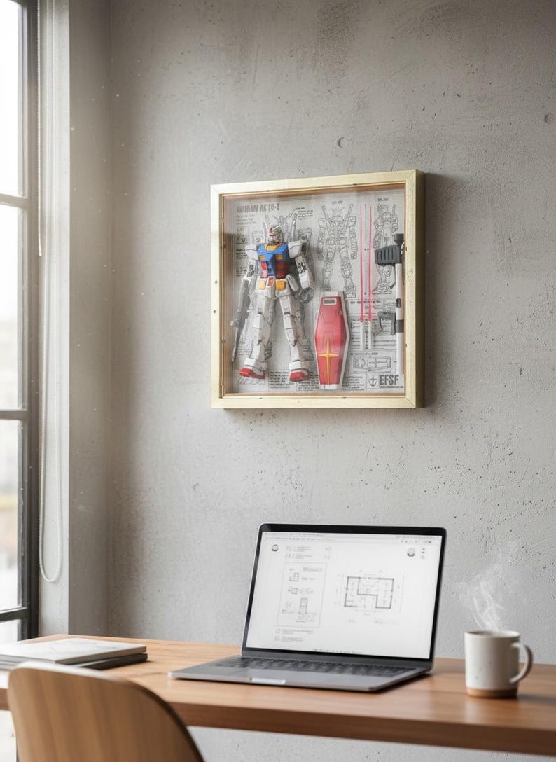 May include: A framed Gundam model kit displayed on a gray wall. The model is in a clear box with a wooden frame, showcasing the robot, shield, and blueprints. A laptop and a mug of coffee sit on a wooden desk in the foreground.