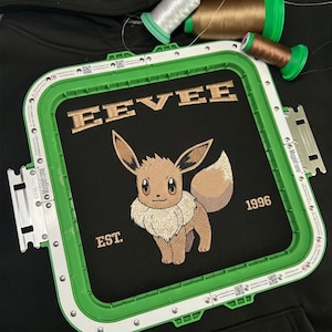 May include: Black fabric with an embroidered design featuring the word "EEVEE" above a cartoon character, with "EST. 1996" below. The design is framed by a green embroidery hoop, with spools of thread in the background.
