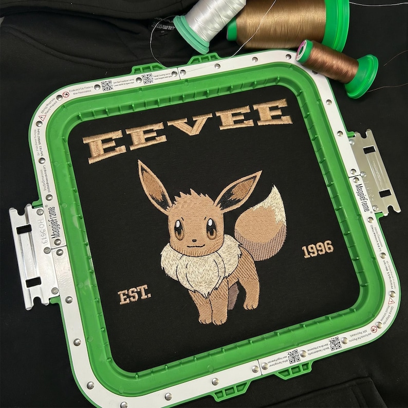 May include: Black fabric with an embroidered design featuring the word "EEVEE" above a cartoon character, with "EST. 1996" below. The design is framed by a green embroidery hoop, with spools of thread in the background.