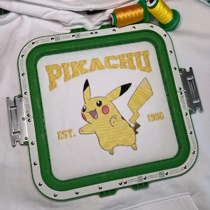 May include: Embroidered Pikachu design in a green embroidery hoop. The design features the word "PIKACHU" above a yellow Pikachu character with the text "EST. 1996" below. Two spools of yellow thread are in the background.