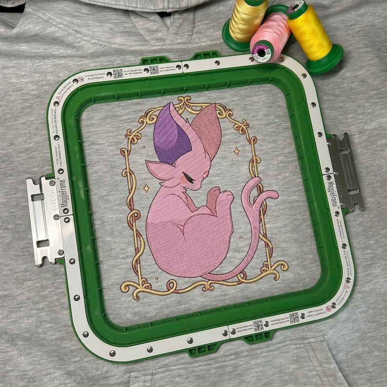 May include: An embroidery hoop with a green frame holds a pink cat design. The cat has purple and pink ears and is inside a gold frame. Three spools of thread in yellow and pink are in the background.