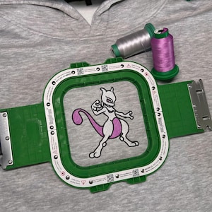 May include: A grey hoodie with an embroidered cartoon character in purple and white, framed by a green embroidery hoop. Two thread spools, silver and purple, are visible. The hoop is labelled "MangoFrame".