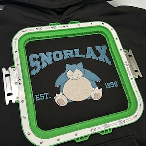 May include: Black hoodie with a green embroidery frame. The hoodie features a blue embroidered design of Snorlax with the text "SNORLAX" above and "EST. 1996" below. The design is centered on the chest.