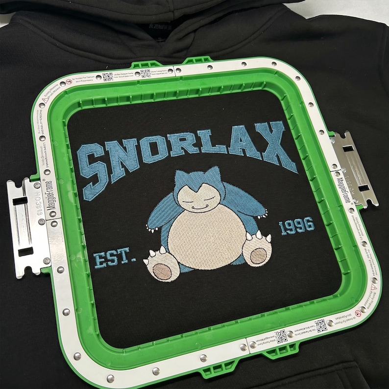 May include: Black hoodie with a green embroidery frame. The hoodie features a blue embroidered design of Snorlax with the text "SNORLAX" above and "EST. 1996" below. The design is centered on the chest.