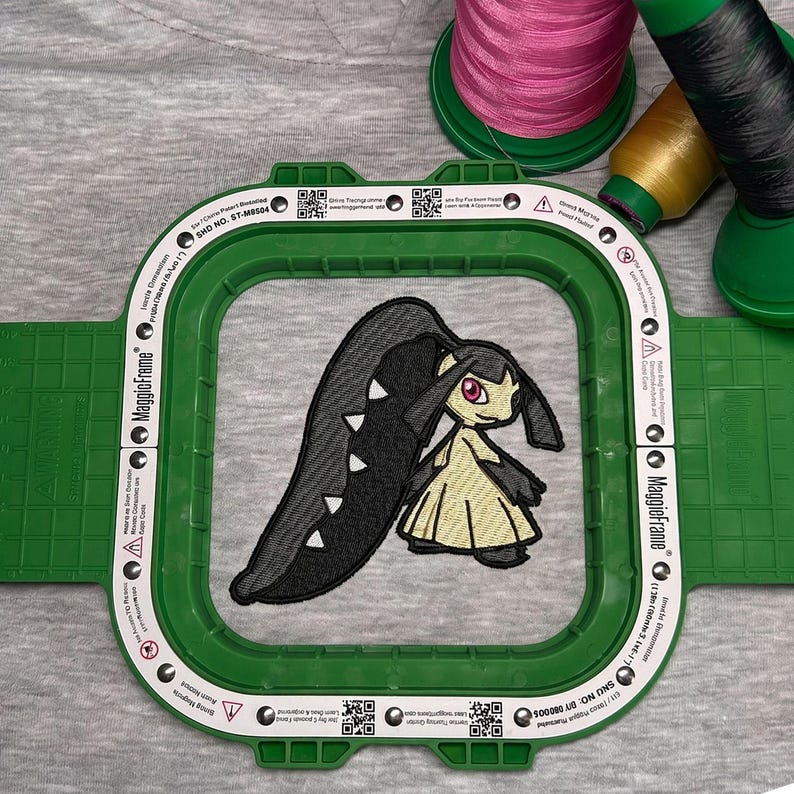 May include: An embroidered patch of a cartoon character in a green embroidery hoop. The character has black and yellow coloring. Pink, yellow, and black thread spools are in the background, indicating a crafting project.
