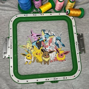 May include: An embroidered design featuring various Pok&eacute;mon characters, including Pikachu, within a green embroidery hoop. Colourful spools of thread in shades of yellow, pink, and green are arranged above the hoop on a grey fabric background.