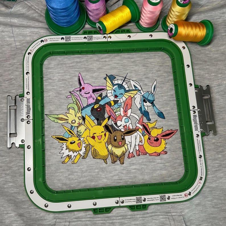 May include: An embroidered design featuring various Pok&eacute;mon characters, including Pikachu, within a green embroidery hoop. Colourful spools of thread in shades of yellow, pink, and green are arranged above the hoop on a grey fabric background.