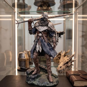 May include: A detailed figurine of a knight in dark blue and silver armour, holding a large sword over their shoulder. The figure stands on a rocky base, with intricate details on the armour and clothing. Other figurines are visible in the background.