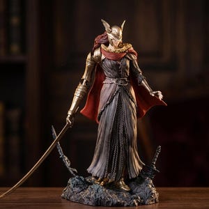 Puede incluir: Figura detallada de una guerrera con un vestido largo y fluido y una armadura dorada, sosteniendo una espada. La figura tiene un casco dorado con detalles de alas y una capa roja. La base es un terreno rocoso.