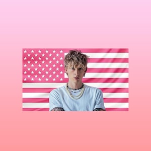 May include: A person wearing a light blue t-shirt and multiple necklaces, posing in front of a pink American flag. The flag has white stars and pink stripes. The background is a gradient of pink.