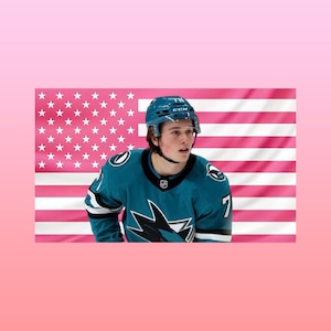 Könnte beinhalten: Ein Eishockeyspieler in einem tealfarbenen Trikot mit der Nummer 7, der einen Helm trägt, steht vor einer rosa amerikanischen Flagge. Das Trikot hat ein Hai-Logo. Der Hintergrund ist ein Rosé-Farbverlauf.