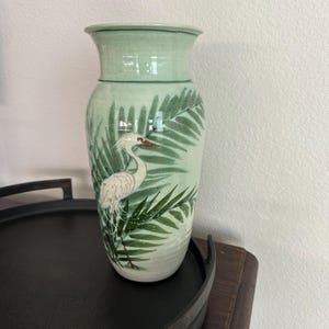 Puede incluir: Un jarrón de cerámica verde claro con un diseño de garza blanca y hojas verdes. El jarrón tiene una parte superior acampanada y un cuerpo cilíndrico. La garza tiene un cuello y un pico largos. Las hojas están pintadas en tonos verdes.