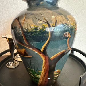Può includere: Un vaso in ceramica dipinto a mano con un corpo largo e un collo stretto. Il vaso presenta un paesaggio con un albero, acqua ed edifici, dipinto in tonalità di blu, marrone e giallo. Il vaso è esposto su un vassoio nero.