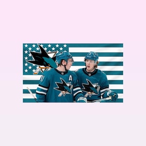Könnte beinhalten: Zwei Eishockeyspieler in tealfarbenen Trikots mit dem Logo der San Jose Sharks, vor einer Flagge mit dem Teamlogo. Die Trikots haben weiße Streifen und den Buchstaben "A". Die Spieler tragen Helme und halten Hockeyschläger.