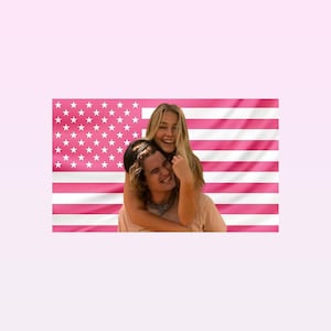 May include: A couple smiling and embracing in front of a pink American flag. The woman is embracing the man from behind, both are smiling. The flag has pink stripes and white stars.