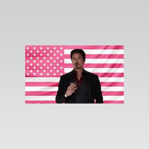 May include: A person with dark hair and a goatee wearing a dark blazer, a dark waistcoat, and a maroon button-down shirt, holding a glass. The background is a flag featuring horizontal stripes and a star field in shades of pink and white.