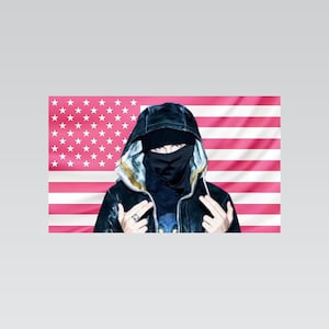 Può includere: Una giacca nera con cappuccio indossata con un berretto nero e una mascherina nera, davanti a una bandiera americana rosa e bianca. La persona fa un gesto con le mani alzando entrambi i medi e indossa un anello d'argento su un dito.