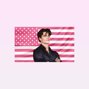 Op de afbeelding: Een persoon in een zwart overhemd glimlacht voor een roze Amerikaanse vlag. De vlag heeft witte sterren in de linkerbovenhoek en roze en witte strepen. De persoon heeft donker haar en kijkt in de camera.