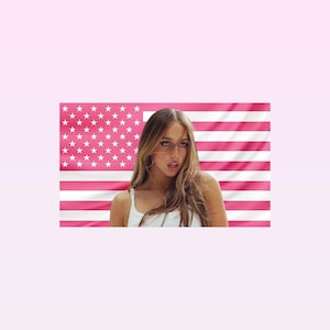 Puede incluir: Una persona con cabello castaño largo que lleva una camiseta sin mangas blanca, posando frente a una bandera estadounidense rosa. La bandera tiene rayas rosas y estrellas blancas. El fondo es de color rosa claro.