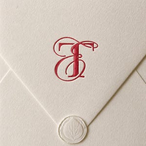 Puede incluir: Sobre de color crema con una letra "E" roja estilizada y un sello de cera transparente. El sello presenta un diseño de hoja. El sobre está doblado y tiene una superficie texturizada.