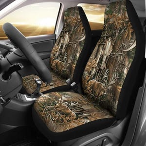Puede incluir: Fundas para asientos de coche con un diseño de camuflaje que representa ciervos y elementos forestales. El diseño utiliza tonos marrones, verdes y beige, creando una escena exterior realista. Los bordes de las fundas son negros.