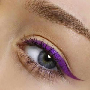 May include: Close-up of an eye with a vibrant purple eyeliner and matching mascara. The eye has a light blue iris and dark eyelashes. The eyebrow is a light brown color.