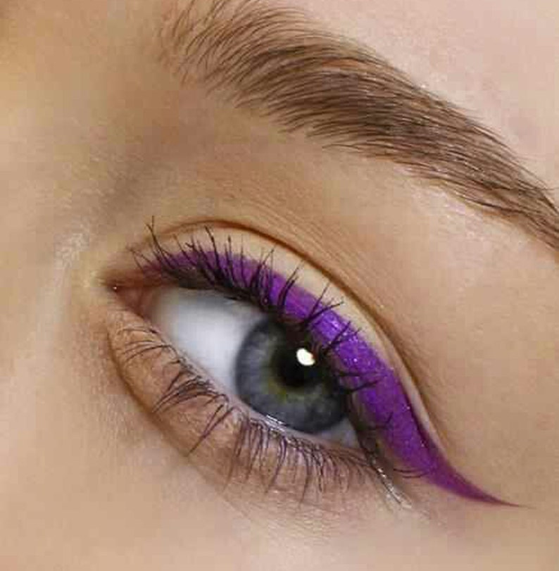 May include: Close-up of an eye with a vibrant purple eyeliner and matching mascara. The eye has a light blue iris and dark eyelashes. The eyebrow is a light brown color.