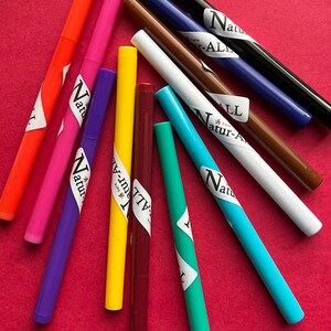 May include: A collection of colorful markers in various shades, including orange, pink, purple, yellow, and blue, arranged on a red surface. Each marker has a white label with the text "Natur-ALL".