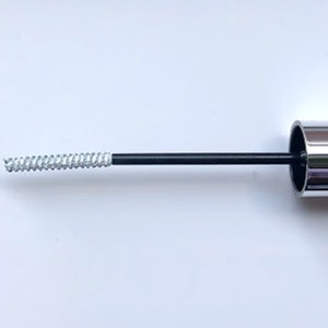 May include: A silver mascara tube with a black wand and a spiral brush. The brush is coated in a light-colored product. The mascara tube has a shiny, metallic finish. The image is set against a white background.