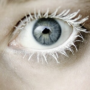 May include: Close-up of a human eye with a light blue iris and white eyelashes. The eye is open, with the pupil appearing black. The skin around the eye is pale, creating a stark contrast with the eye's colors. The image has a soft focus.