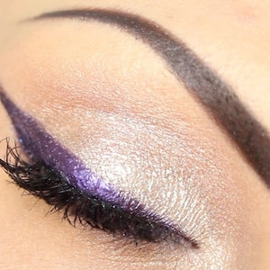 May include: Close-up of an eye with makeup. The eyelid is covered in a shimmery, light-colored eyeshadow. A bold, purple eyeliner is applied along the upper lash line. The eyelashes are coated in black mascara, and the eyebrow is dark.