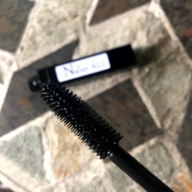 May include: Close-up of a black mascara wand with a bristled applicator. The mascara tube is black with a white label that reads "Natur ALL". The background is a stone tile surface.