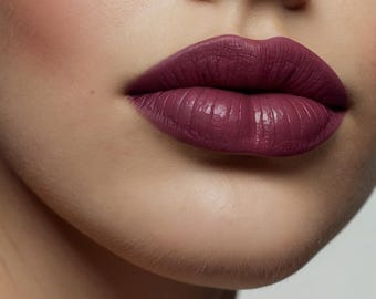 May include: Close-up of a person's lips, fully covered in a deep plum lipstick. The lipstick has a matte finish and accentuates the natural lip lines. The skin around the lips is smooth and fair, creating a striking contrast with the bold lip color.