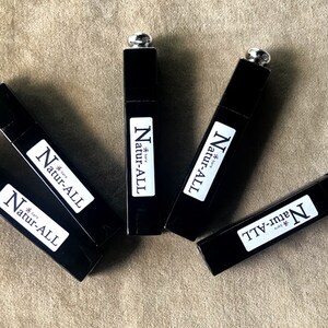 May include: An overhead shot of five black rectangular tubes with silver caps. Each tube has a white label with the text "Natur-ALL" in a bold, sans-serif font. The tubes are arranged on a neutral-toned surface.