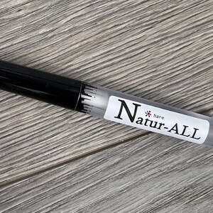 May include: A clear, cylindrical tube with a black cap. The tube has a white label with the text "Natur-ALL bare". The product appears to be a cosmetic item, possibly a lip gloss or a similar beauty product. The background is a light gray wooden surface.