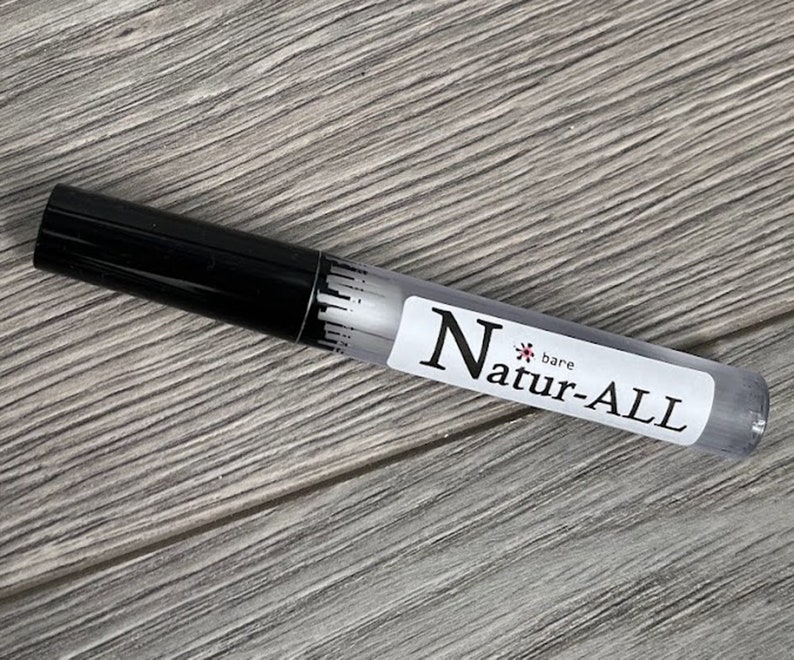 May include: A clear, cylindrical tube with a black cap. The tube has a white label with the text "Natur-ALL bare". The product appears to be a cosmetic item, possibly a lip gloss or a similar beauty product. The background is a light gray wooden surface.