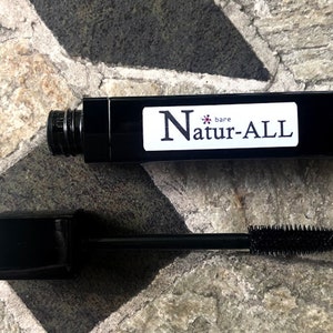 May include: Black mascara tube with the text "bare Natur-ALL" on a white label. The mascara wand is extended, showing the brush. The mascara tube and wand are on a stone surface.