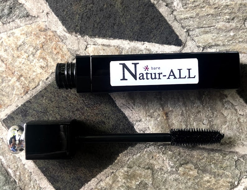 May include: Black mascara tube with the text "bare Natur-ALL" on a white label. The mascara wand is extended, showing the brush. The mascara tube and wand are on a stone surface.