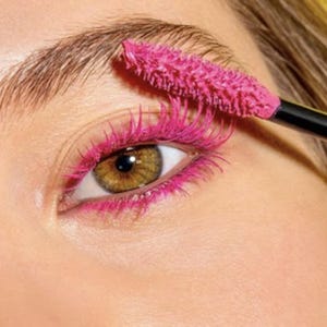May include: Close-up of an eye with bright pink mascara applied to the upper and lower lashes. A black mascara wand is visible, applying the vivid colour. The eye has a light brown iris and is set against a pale skin tone.