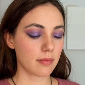 May include: Close-up of a person's face with eye makeup. The eyelids are covered in a shimmering purple eyeshadow, with a sharp, winged eyeliner. The lips are a soft pink, and the skin has a natural, healthy glow.