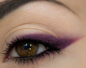 May include: Close-up of an eye with brown iris and dark eyelashes. The eyelid is adorned with a deep purple eyeshadow, extending into a sharp wing. The eyebrow is visible above the eye, and the skin has a smooth texture.
