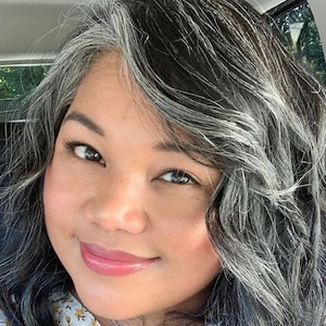 May include: A close-up portrait of a person with dark hair, streaked with silver and gray. The person has brown eyes and is smiling slightly, with a light pink lip color. The background is blurred, suggesting an outdoor setting.