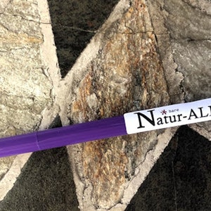 May include: A purple pen with the text "Natur-ALL" on a white label. The pen is lying on a stone surface with a diamond-shaped pattern. The pen's cap is a darker shade of purple.