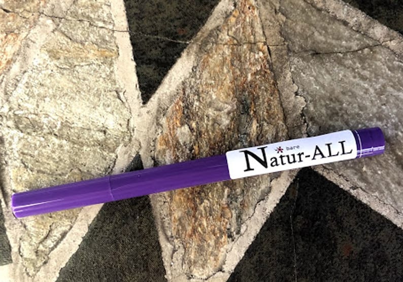 May include: A purple pen with the text "Natur-ALL" on a white label. The pen is lying on a stone surface with a diamond-shaped pattern. The pen's cap is a darker shade of purple.