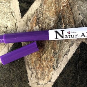 May include: A purple Natur-ALL bare pen with a black tip and a cap lying next to it. The pen has a white label with the brand name printed in black. The pen is on a textured stone surface.