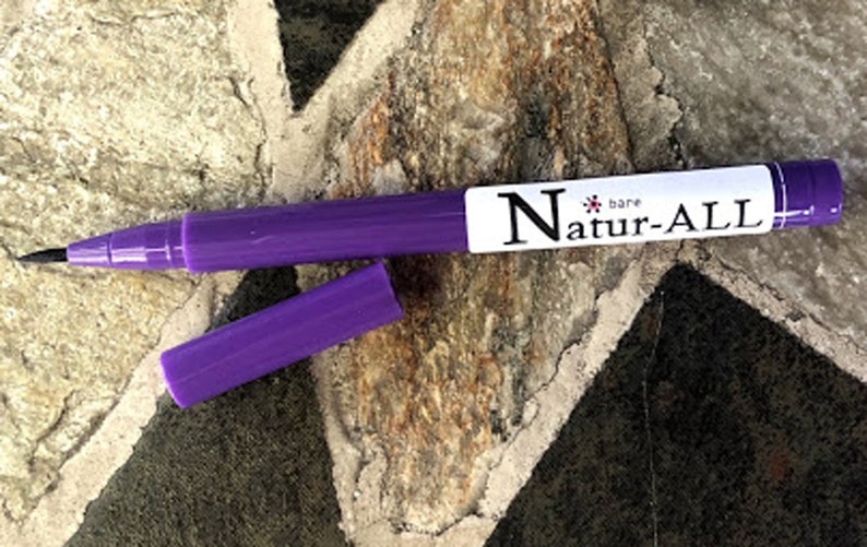 May include: A purple Natur-ALL bare pen with a black tip and a cap lying next to it. The pen has a white label with the brand name printed in black. The pen is on a textured stone surface.